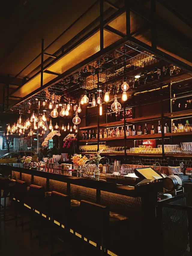 A long bar lit by warm hanging bulbs, bottles stacked to the ceiling.