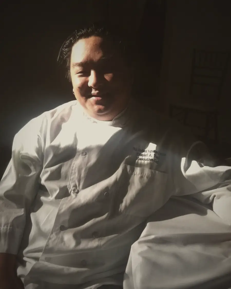 Chef Daniel Vogel in his whites, smiling, half-lit by a shaft of afternoon sun.