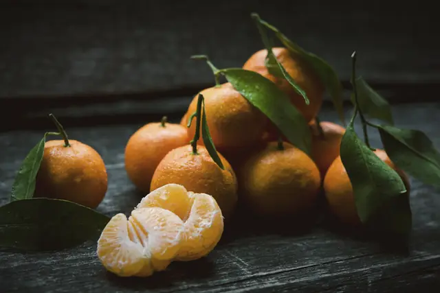 Mandarins with their leaves on a dark wooden table, one peeled.