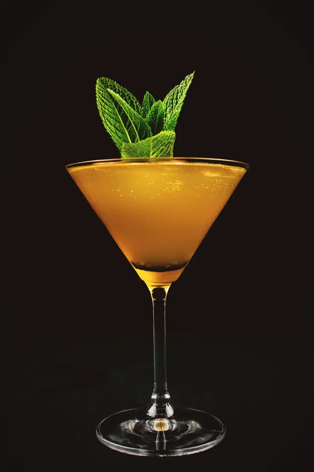 An amber cocktail in a coupe with a sprig of mint, against black.