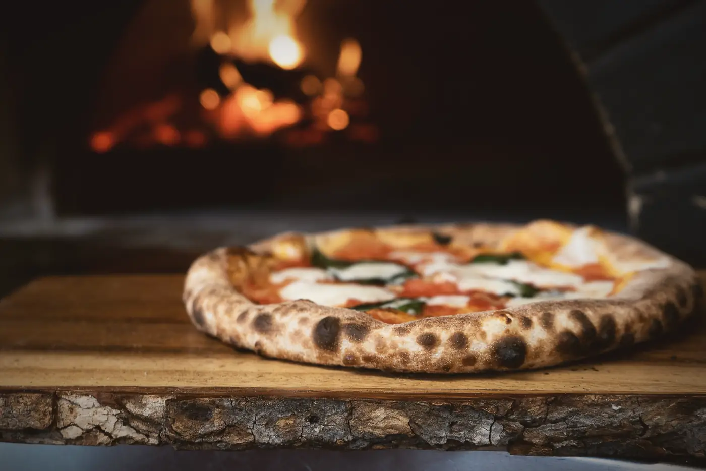 A blistered pizza resting on a wooden board at the mouth of the wood-fired oven, flames behind it.